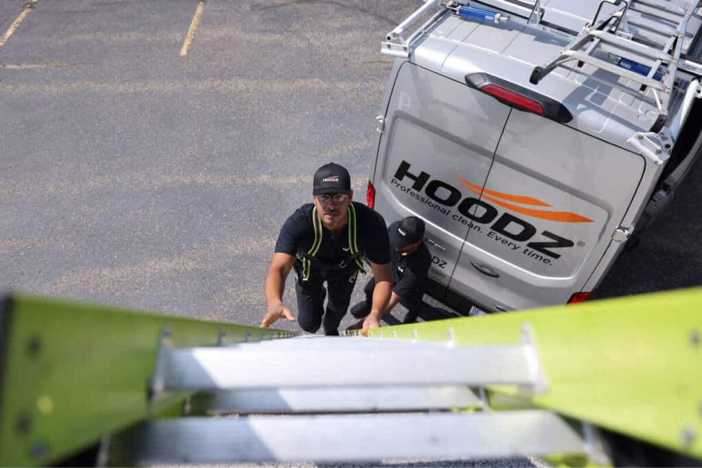Two workers climb a ladder against a van marked "Hoodz Professional Clean." The ground is paved, and the van's license plate is visible.