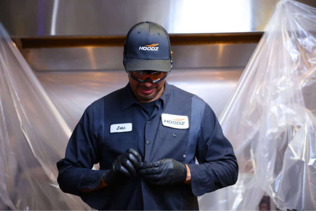 A person wearing a dark uniform and cap, labeled "Hoodz," stands in front of a covered kitchen area, adjusting their gloves.